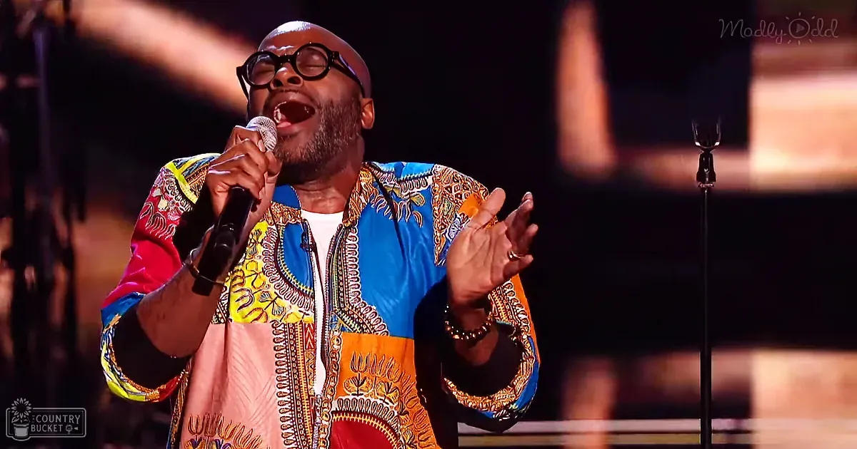 A middle-aged male singer performing on a dimly lit stage, wearing a colorful traditional African print garment with vibrant blue, yellow, orange, and red hues.