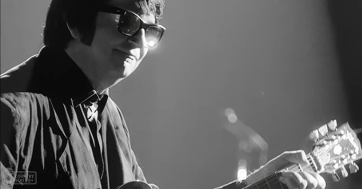 A man wearing glasses plays a guitar, smiling, in a black-and-white image.