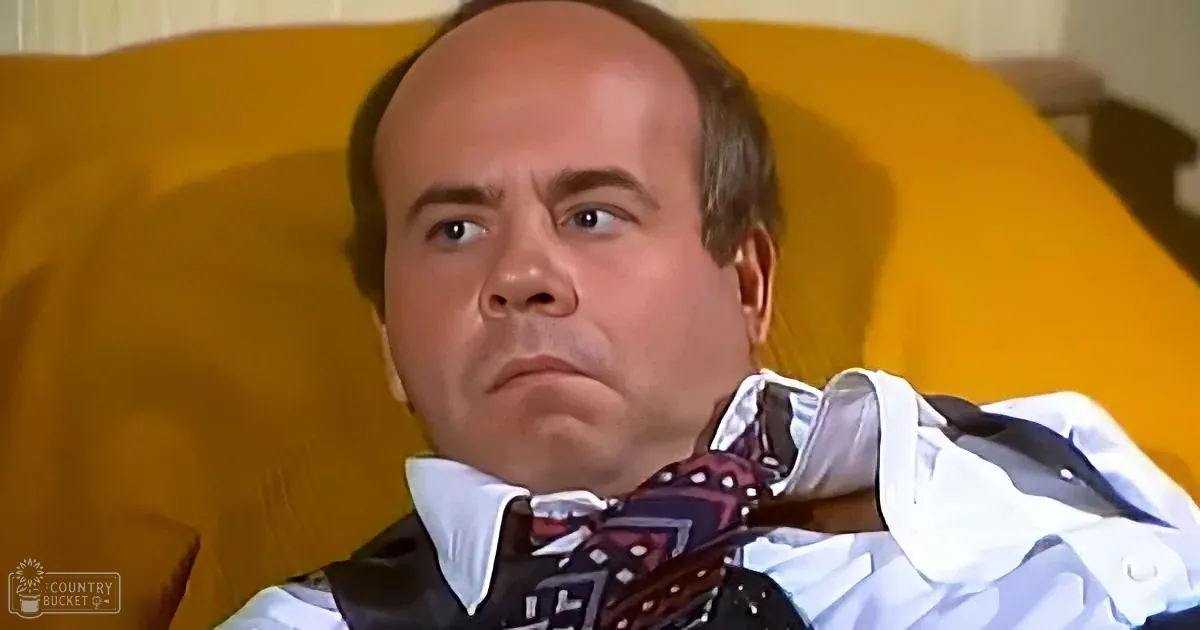A man in a white shirt and bow tie sits on a yellow couch, looking slightly to the side with a serious expression.