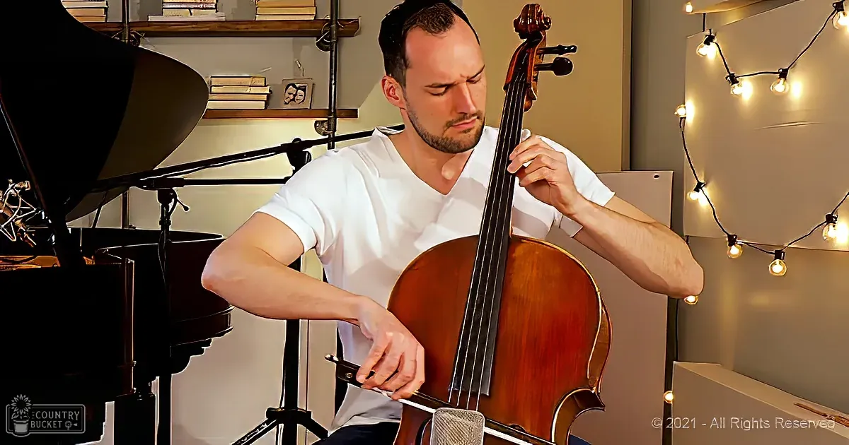 A man in a white shirt plays a cello in a cozy room with string lights and a piano.