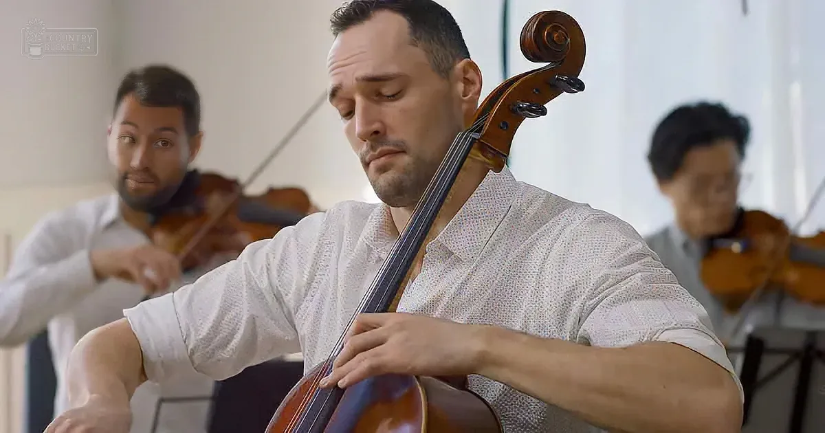 A man in a white shirt plays a cello with two violinists in the background.