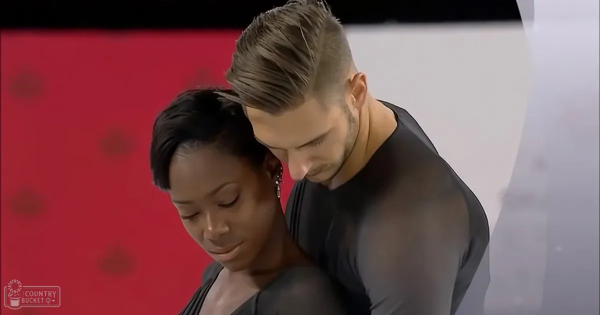 Young adult female dancer in a black silk dress performs alongside another young adult male dancer wearing a black sleeveless top, with a red backdrop suggesting a dance studio or stage setup.