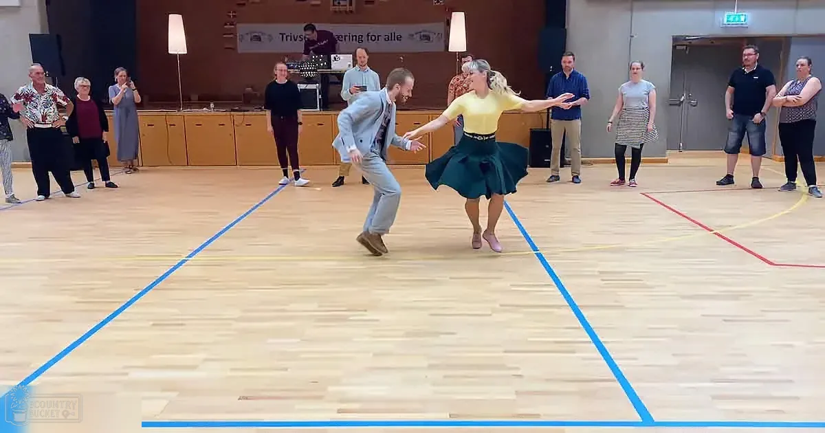 A man and woman dance in a gymnasium, surrounded by onlookers, with a banner in the background.