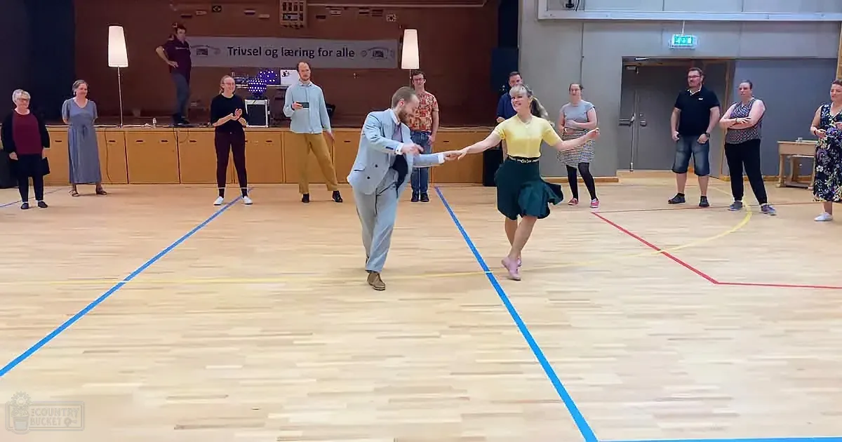 A man and woman dance in a gymnasium, surrounded by onlookers, with a banner in the background.