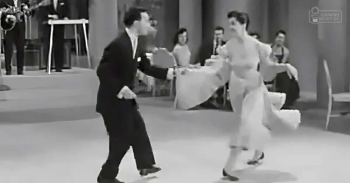A man and woman are dancing in a black-and-white scene, with the man leading and the woman following gracefully.