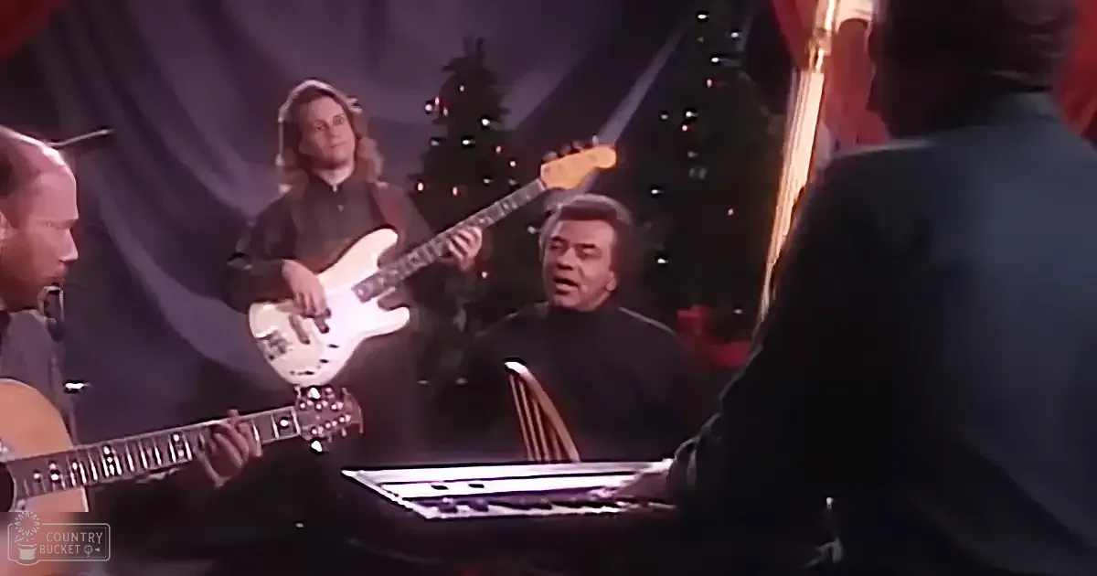A musician plays guitar while another sings in front of a Christmas tree, with a keyboardist and guitarist in the foreground.