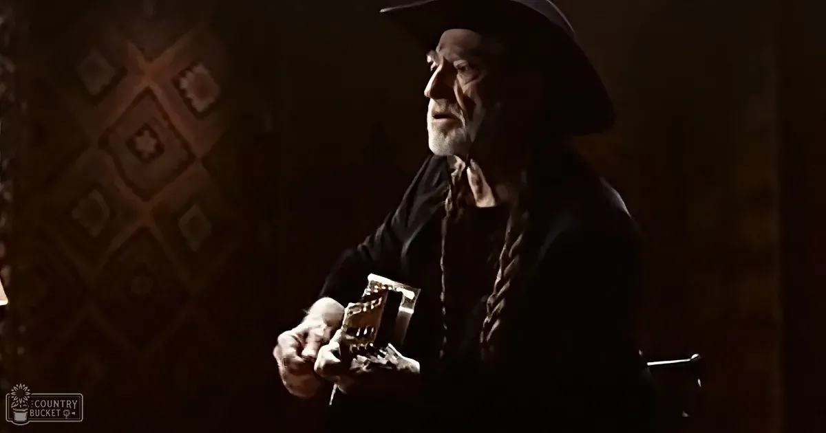 An older man in a cowboy hat holds a guitar, wearing a dark shirt and tie, in a dimly lit room with patterned wallpaper.