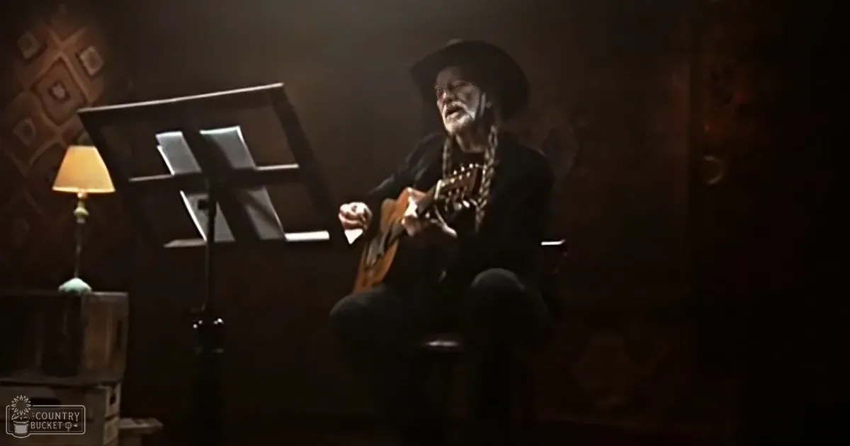 An older man in a cowboy hat plays guitar, surrounded by rustic decor and a lamp.