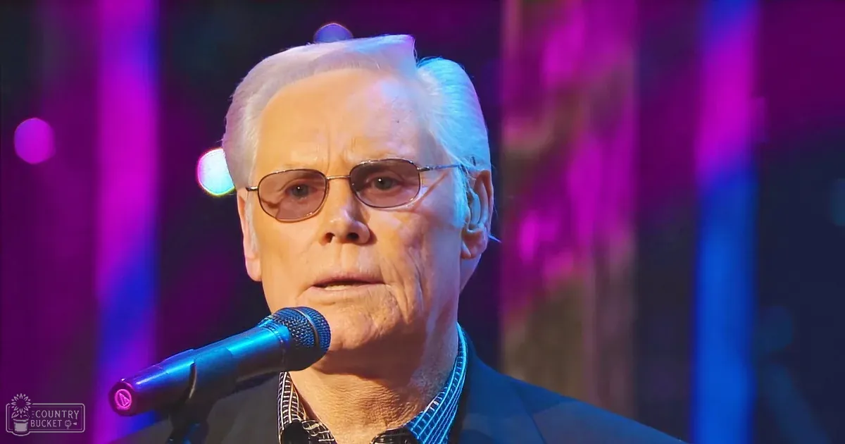 A middle-aged adult male wearing a formal dark suit and white shirt, performing on an indoor stage with festive lighting.