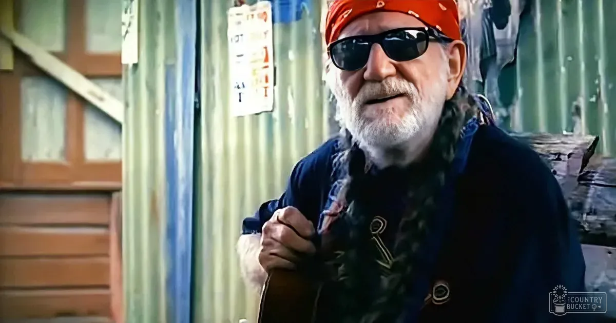 An older man with a white beard and sunglasses, wearing a red bandana, holds a guitar in front of a corrugated metal wall.