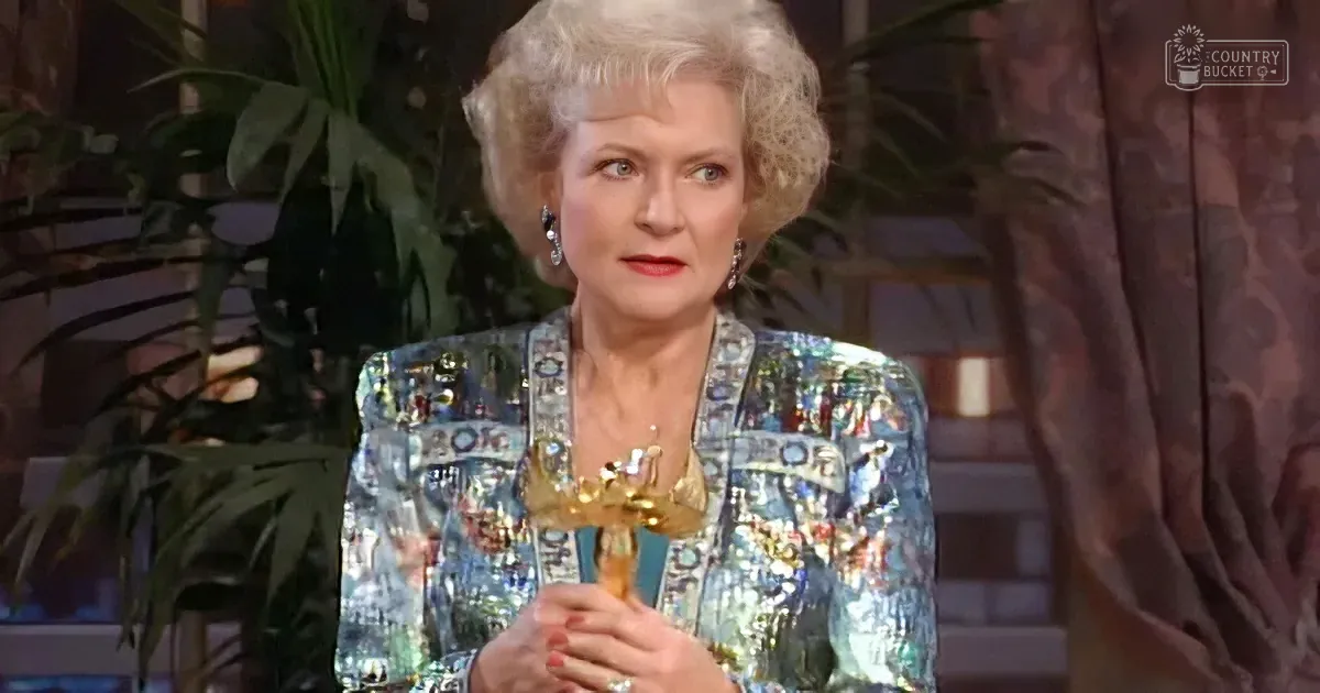 An older woman with blonde hair, wearing a colorful patterned jacket, holds a trophy in a studio setting with plants in...