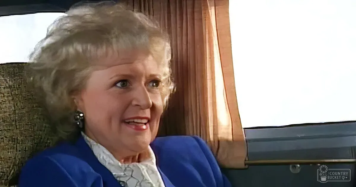 An older woman with curly gray hair, wearing a blue jacket and white shirt, sits in a car with a beige interior and a...