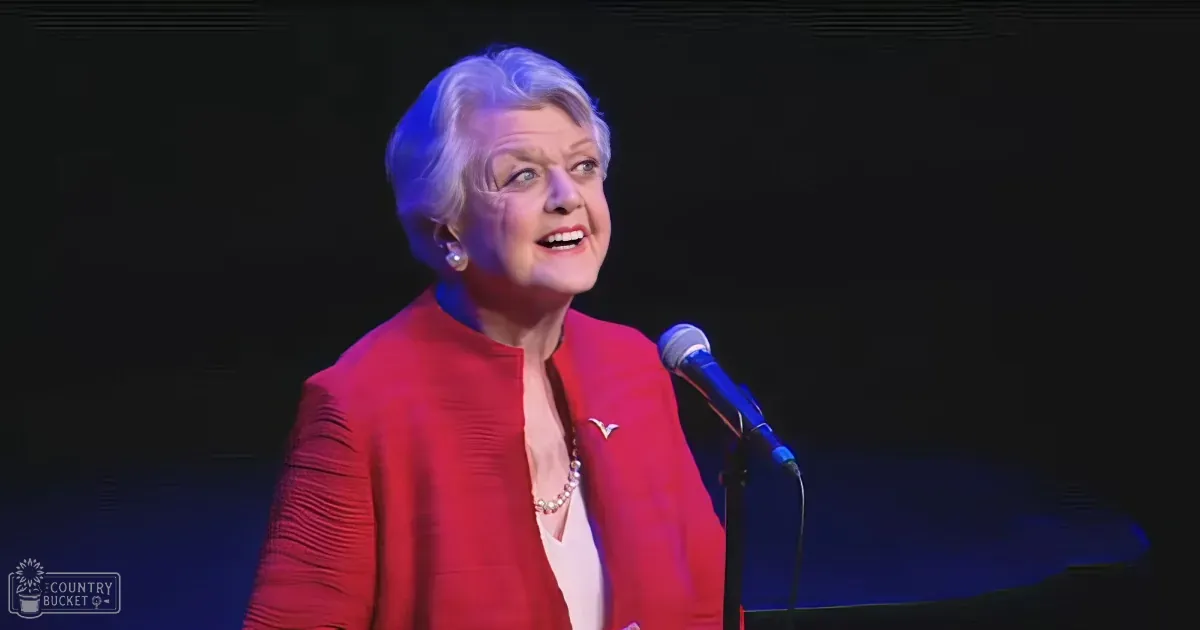 An older woman in a red outfit speaks into a microphone on stage.