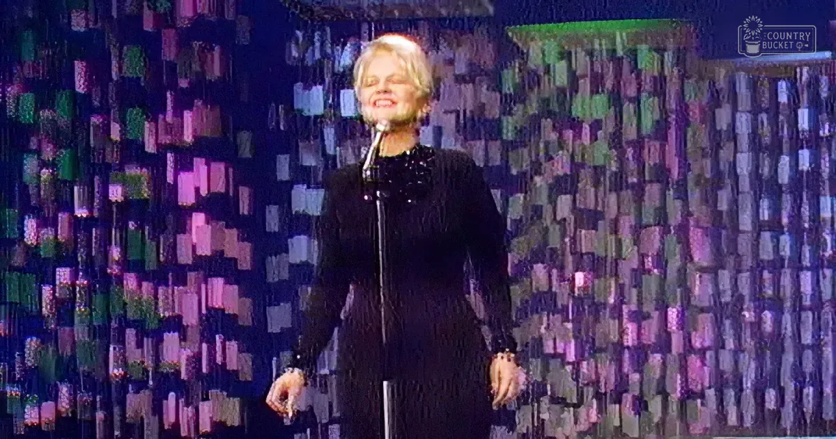 A person in a black outfit stands on stage with a microphone, surrounded by a colorful, patterned backdrop.