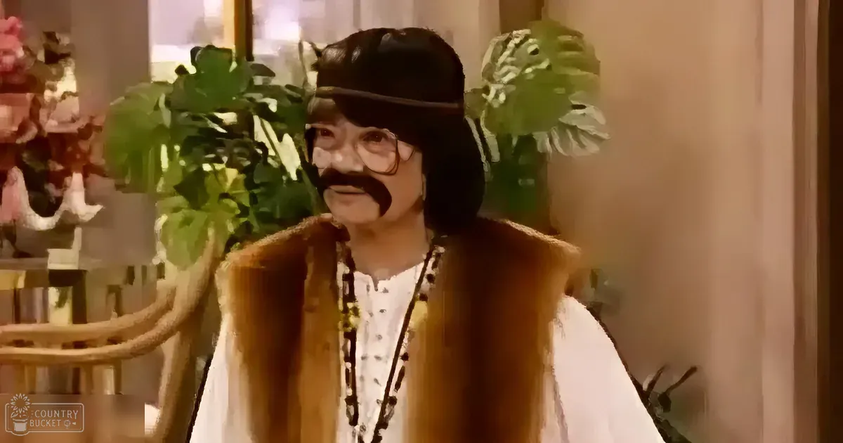 A person in a brown vest, white shirt, and black hat with a mustache stands in a room with plants and flowers.