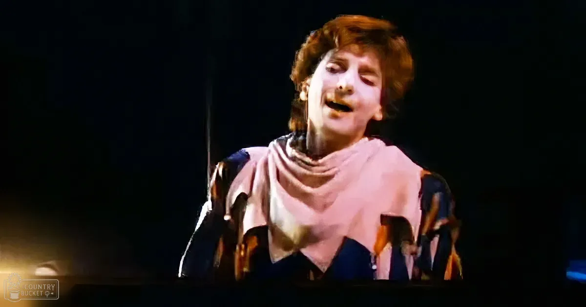 A person in a colorful outfit with a pink scarf is singing on stage, illuminated by stage lights.