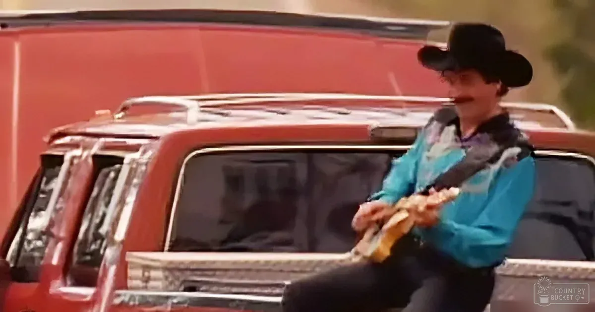 A person in a cowboy hat and blue shirt plays a guitar in front of a red truck.
