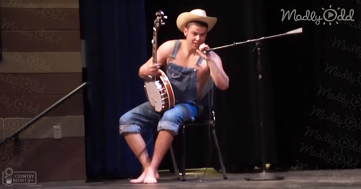 A young adult male singer in denim overalls and a cowboy hat performs on stage holding a banjo, with a microphone stand and dark curtain backdrop.