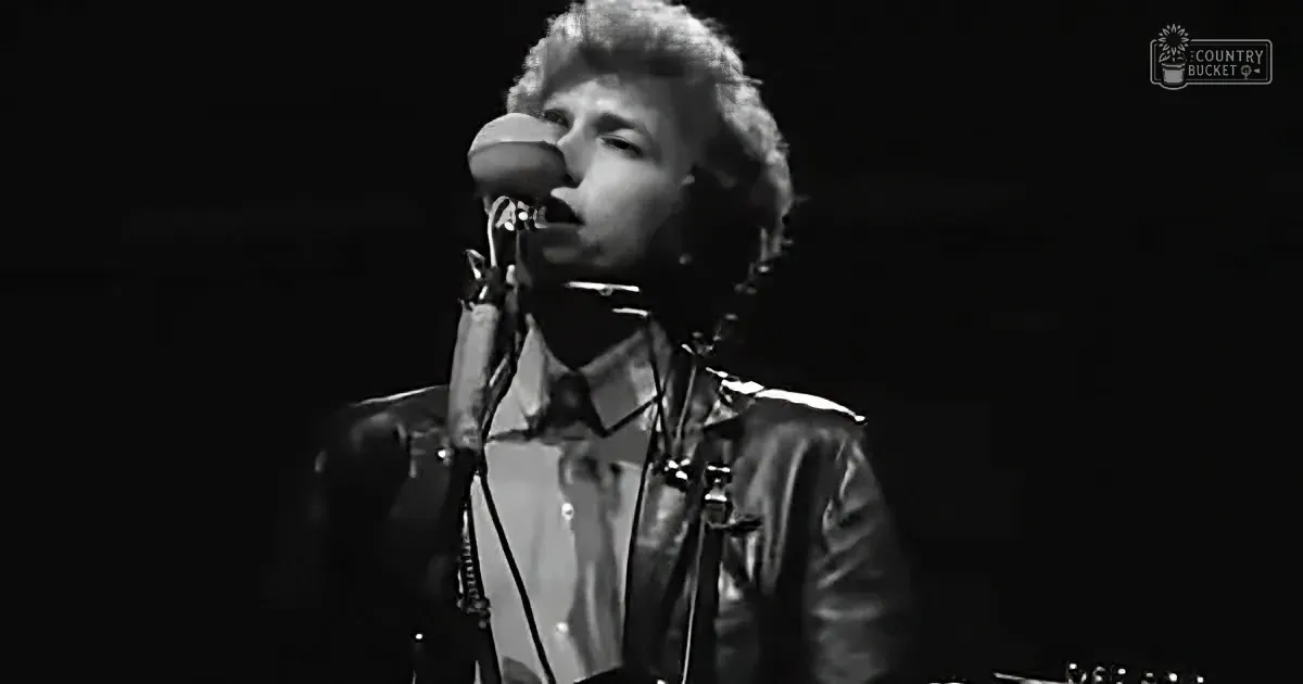 A person in a leather jacket sings into a microphone on stage.