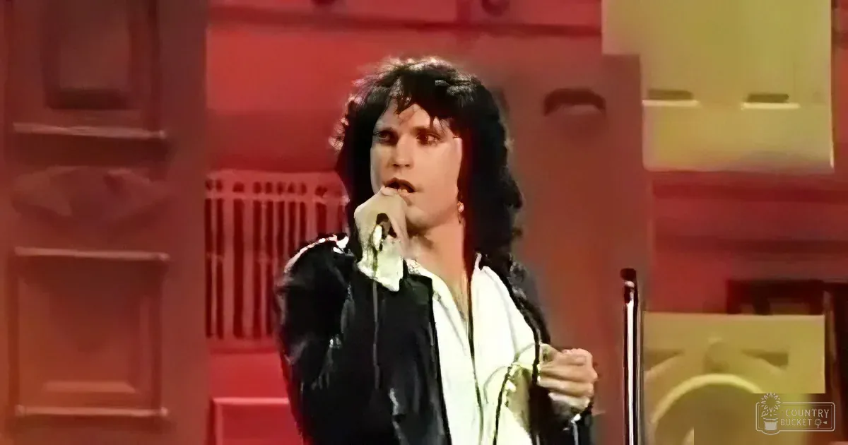 A person with long hair, wearing a black jacket and white shirt, sings into a microphone on stage.