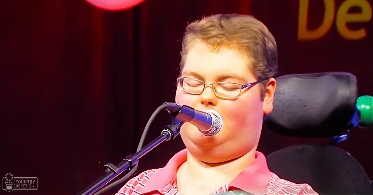 A young adult male singer wearing a green t-shirt with red text, holding a vintage-style microphone in an indoor setting with warm lighting and textured wallpaper.