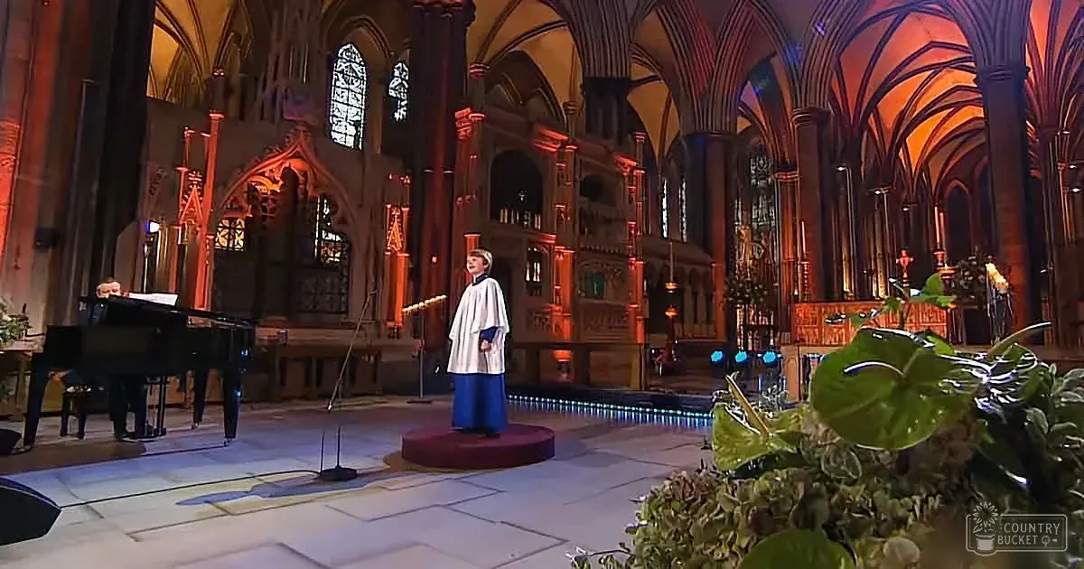 A person in white attire stands on a stage in a grand church, with a pianist and microphone nearby.