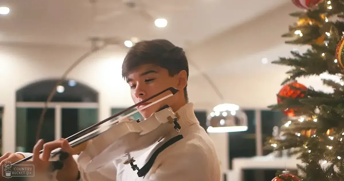 A person in a white shirt plays the violin near a decorated Christmas tree.