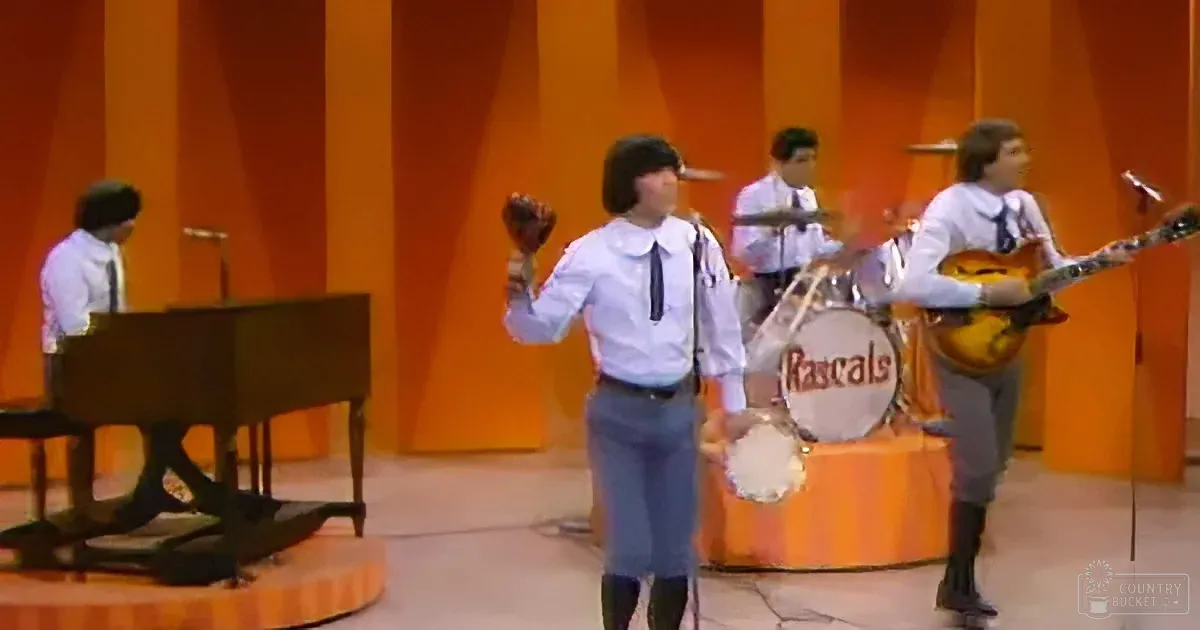 A person in a white shirt and tie plays a tambourine on stage with a band.