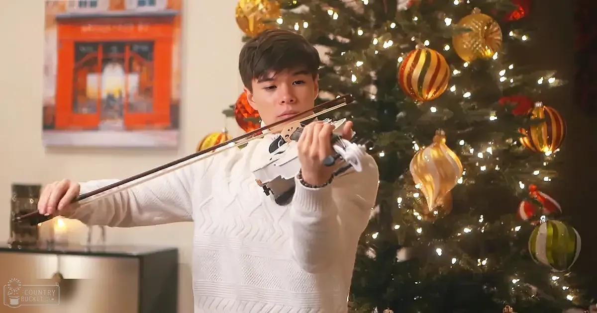 A person in a white sweater plays the violin in front of a decorated Christmas tree.
