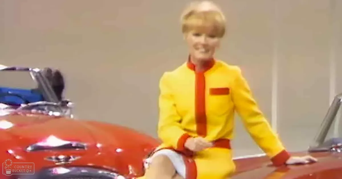 A person in a yellow suit sits on a red car, smiling, with a blurred background.