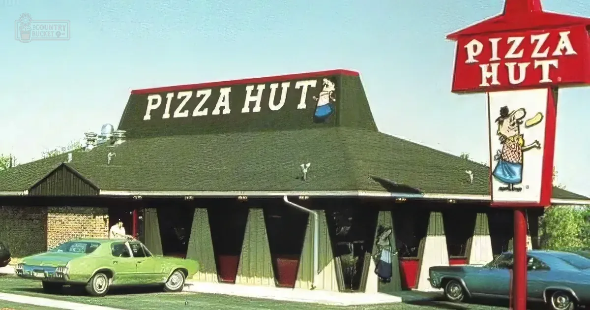 Pizza Hut restaurant with iconic red sign and cartoon character, vintage cars parked, green roof, and brick facade.