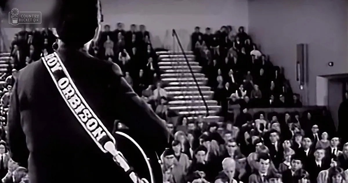 Roy Orbison, wearing a sash, stands on stage with a microphone, facing a crowd in a large auditorium.