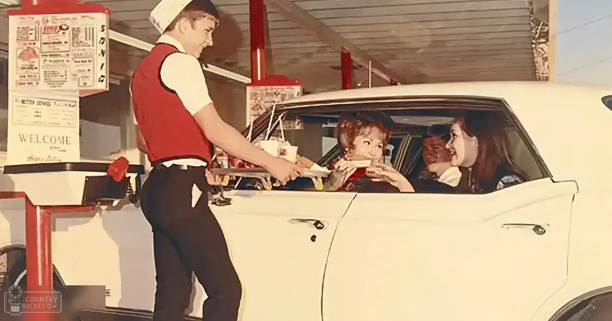 A server in a red vest hands food to a woman in a car at a drive-in.
