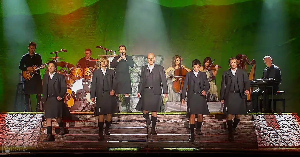 Six men in kilts perform on stage with a band and harpist behind them.