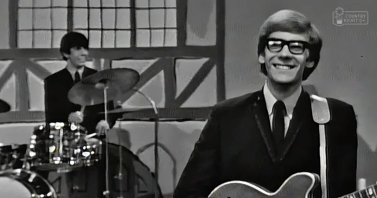 A smiling man in a suit holds a guitar, with a drummer in the background, both in a studio setting.