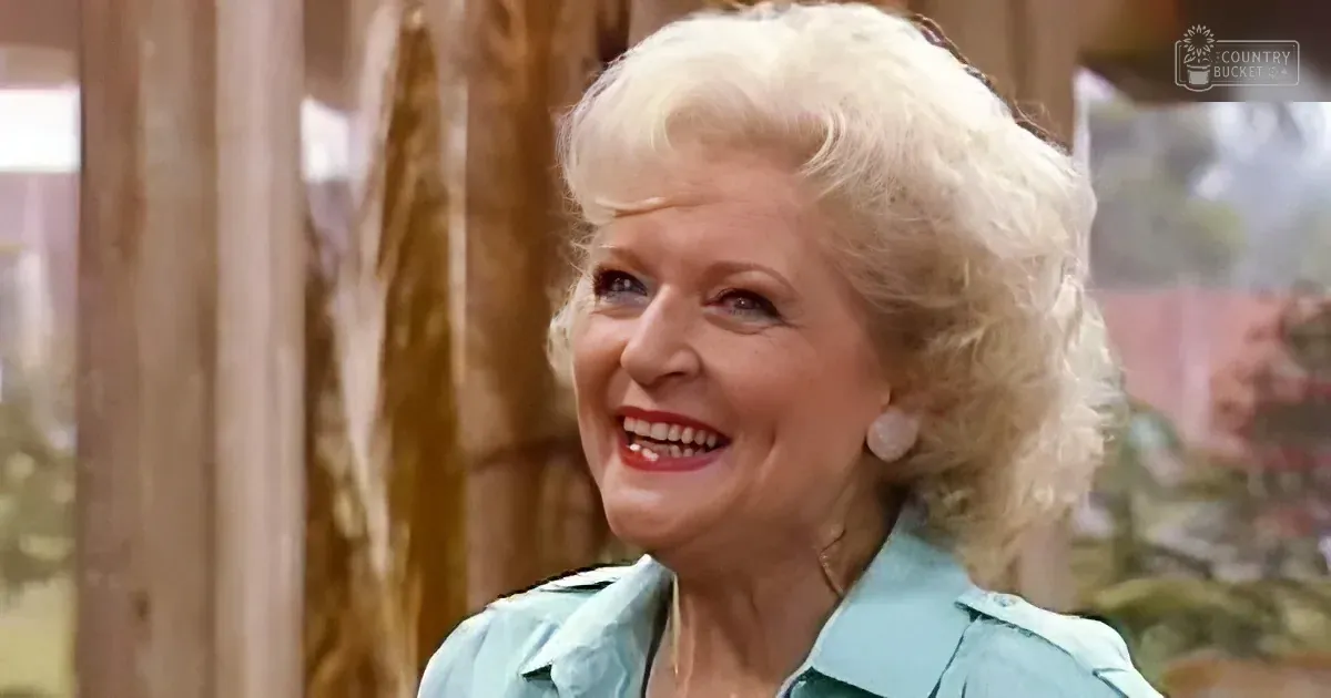 A smiling woman with blonde hair, wearing a light blue shirt, stands in a room with wooden elements and a blurred background.