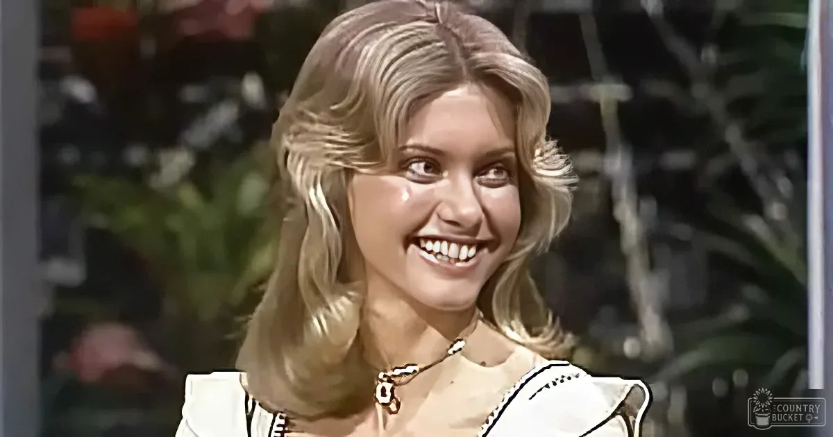 A smiling woman with blonde hair, wearing a white top and a necklace, stands in front of a blurred background with greenery.
