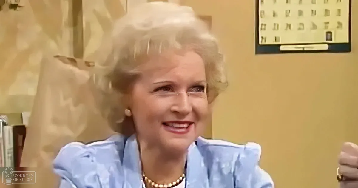A smiling woman with blonde hair, wearing a blue dress and pearl necklace, sits in a room with a calendar on the wall.