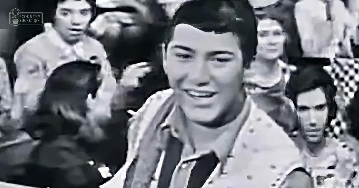 A smiling young man in a patterned shirt, surrounded by a crowd of people, in a black-and-white image.