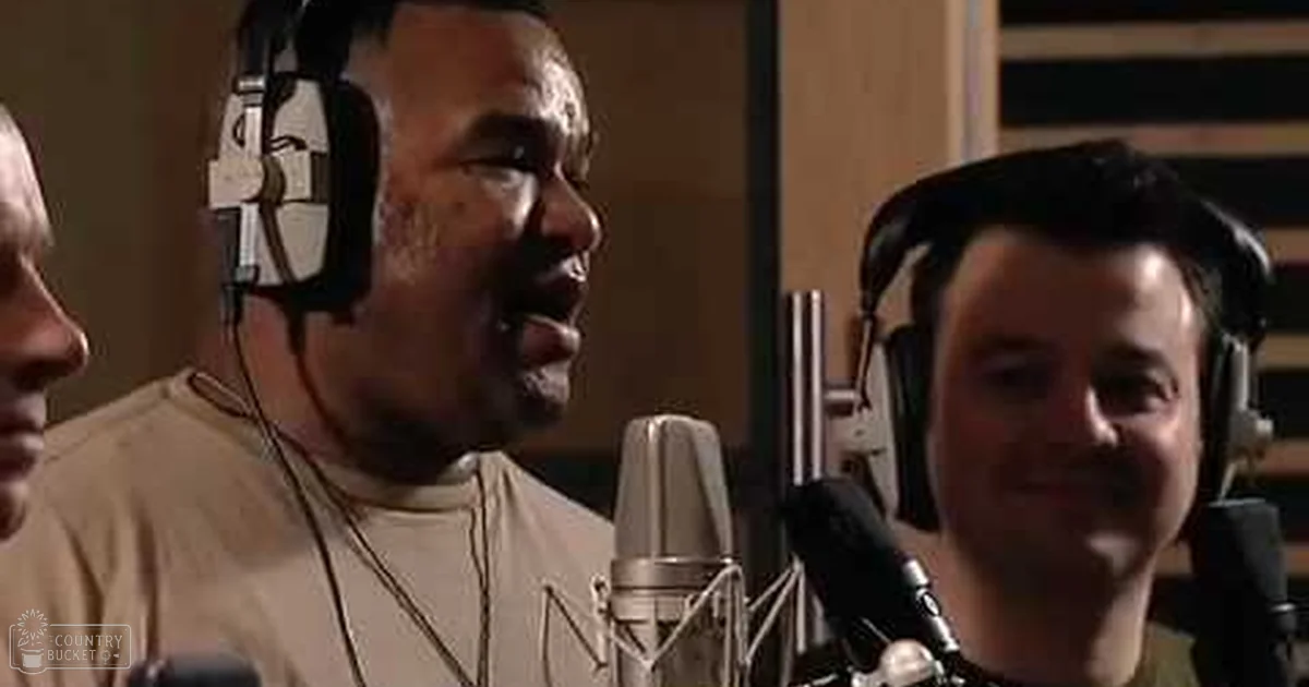 A man wearing a white t-shirt with a visible necklace chain, singing in an indoor professional recording studio setup.