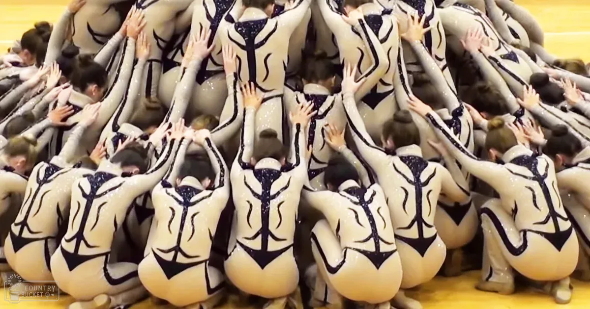 Young adult female gymnasts perform a synchronized routine in an indoor sports arena with yellow flooring, under artificial lighting.