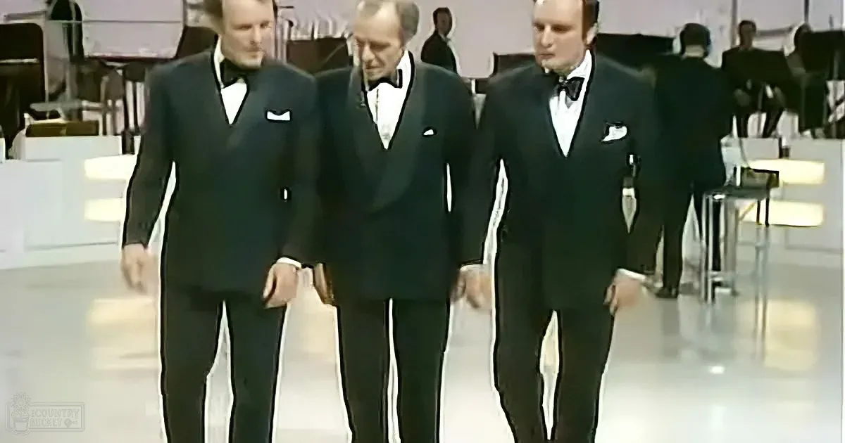 Three men in black tuxedos walk together in a formal setting, possibly a performance or event.