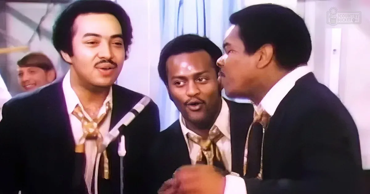 Three men in suits sing into microphones, one holding a glass, with a smiling background figure.