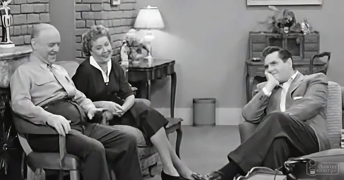 Three people in a living room, two men and a woman, seated and smiling, with a lamp and furniture in the background.