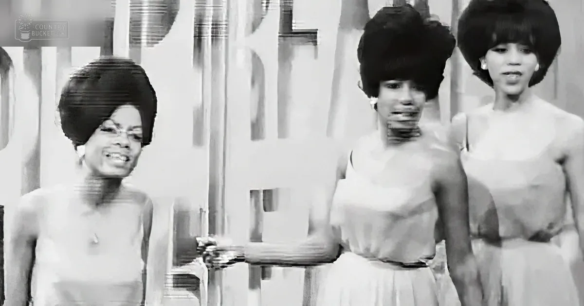 Three women in 1960s attire, one holding a microphone, standing in front of a backdrop.