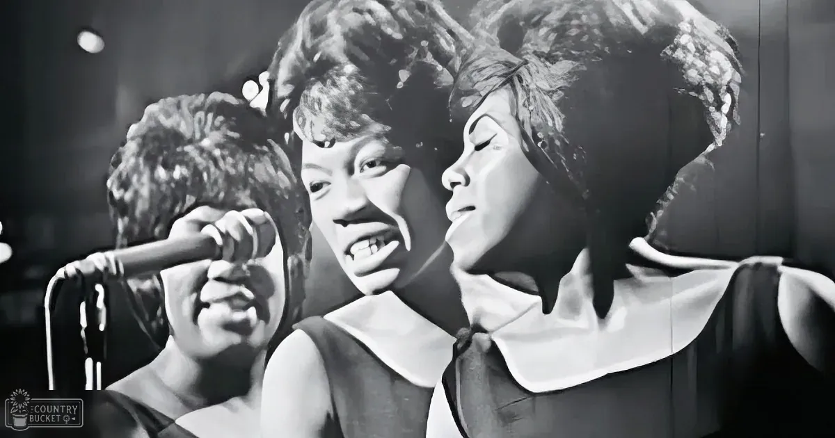 Three women in black dresses sing passionately into a microphone, their expressions intense.