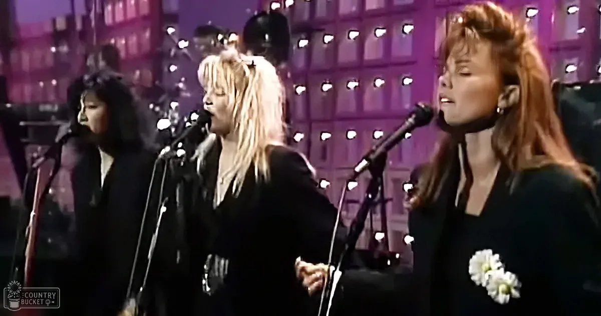 Three women sing into microphones on stage, wearing black, with a cityscape backdrop.