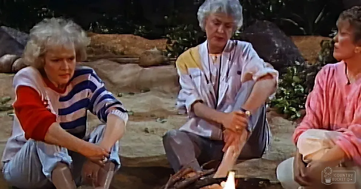 Three women sit outdoors, one in a striped shirt, another in white, and the third in pink, around a fire.