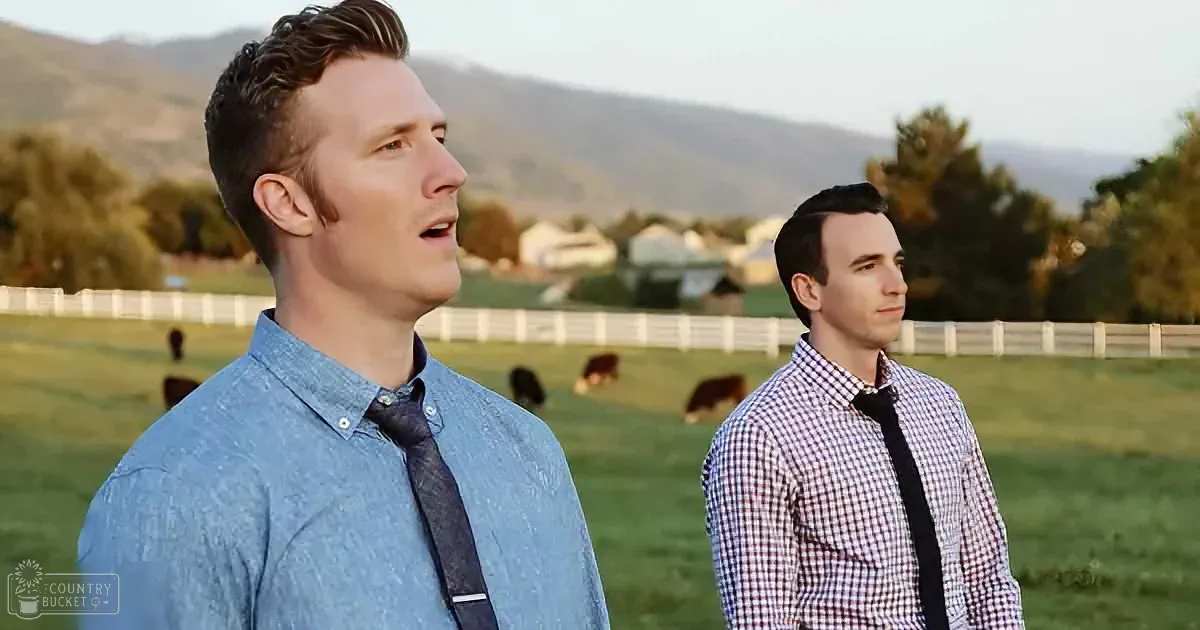 Two men in shirts and ties stand in a grassy field with cows and mountains in the background.
