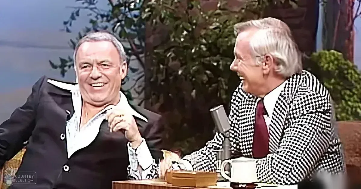 Two men in suits are laughing, one in a black suit and the other in a checkered suit, seated at a table with a microphone...